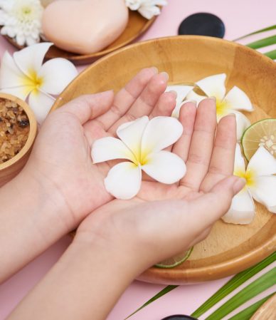 Woman soaking her hands in bowl of water and flowers, Spa treatment and product for female feet and hand spa, massage pebble, perfumed flowers water and candles, Relaxation. Flat lay. top view.
